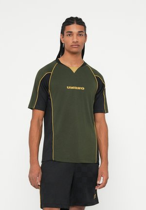 Jaunas vyras su kasytėmis, vilkintis alyvuogių žalios ir juodos spalvos "Umbro" sportinį marškinėlį ir juodas kelnes su subtiliu languotu raštu.