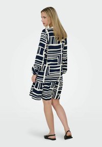 Granatowa i biała sukienka w wzory z długimi rękawami, z abstrakcyjnymi prostokątnymi kształtami i marszczonym dołem, uszyta z lekkiego materiału.