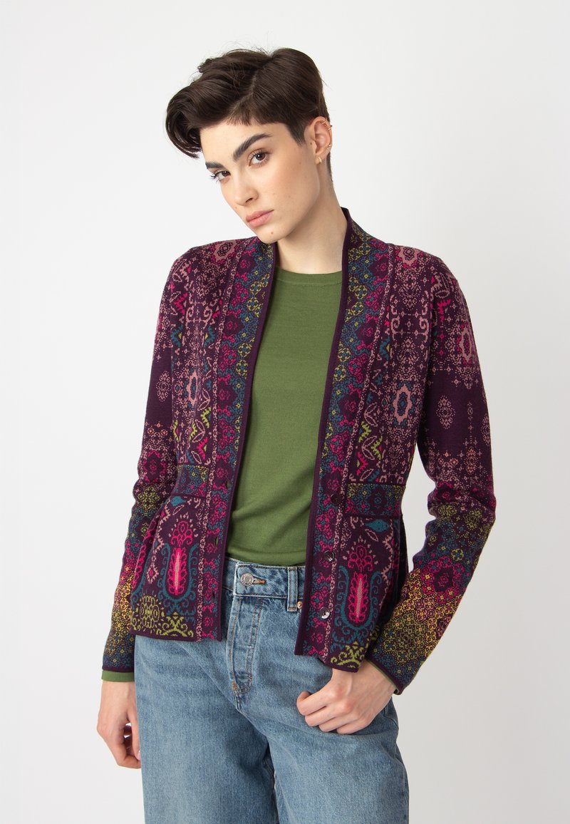 Gilet violet à motifs avec des designs complexes en rose, bleu et vert. Présente des manches longues et une coupe décontractée, superposé sur un haut vert.