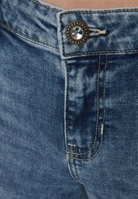 Jeans in denim di colore blu sbiadito, con bottone metallico rotondo decorato con gemma trasparente, cuciture in evidenza e una leggera texture invecchiata.