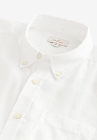 Chemise en lin blanc à boutons, avec un col et des boutons perlés, ainsi qu'une poche sur la poitrine gauche. L'étiquette indique "NEXT SIGNATURE M."
