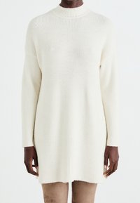 Pull en tricot couleur crème avec un col haut, texture côtelée et manches longues, présentant une coupe légèrement ample et un ourlet asymétrique.