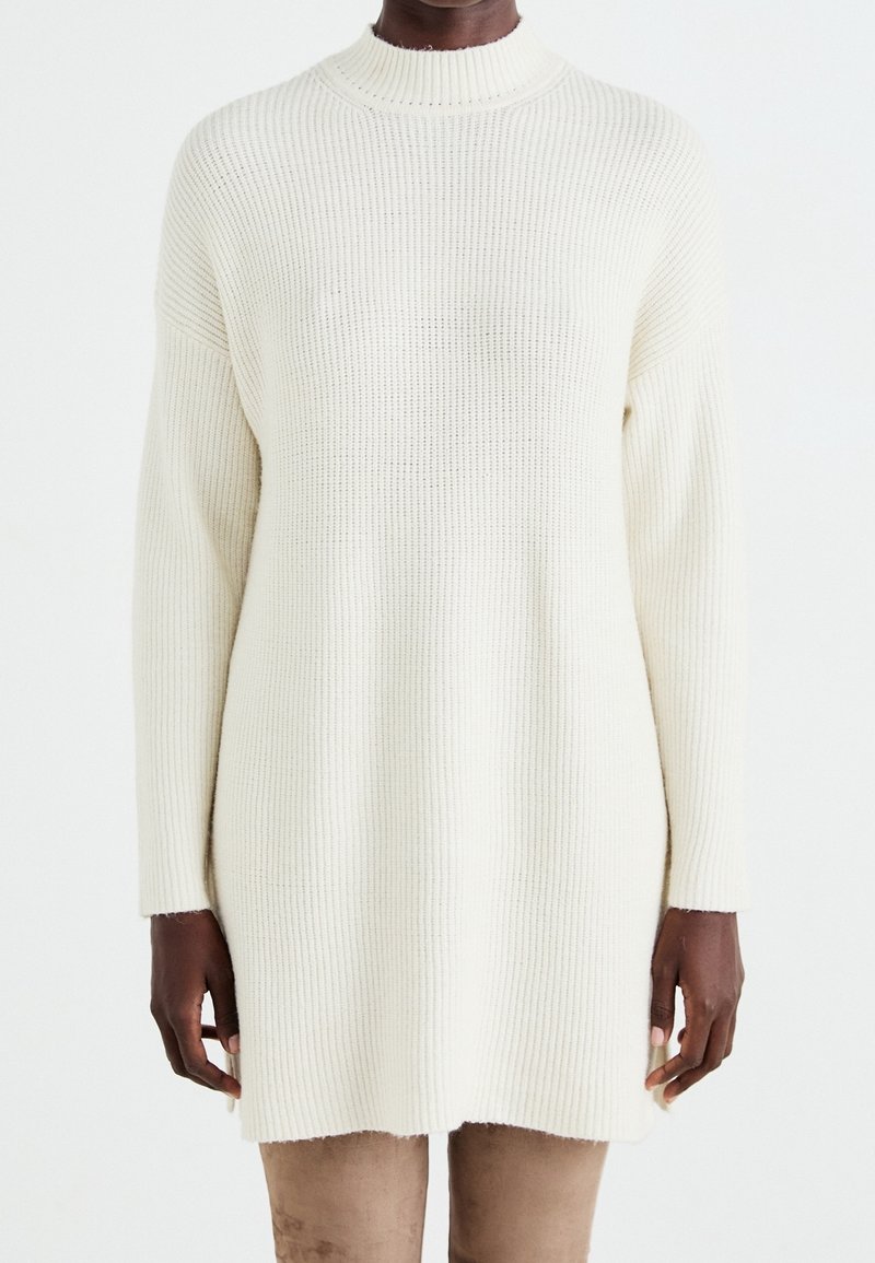 Pull en tricot couleur crème avec un col haut, texture côtelée et manches longues, présentant une coupe légèrement ample et un ourlet asymétrique.