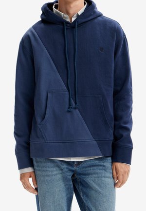 Man wearing dark blue pullover hoodie with a diagonal lighter blue panel, front pocket, and drawstrings, paired with blue jeans.