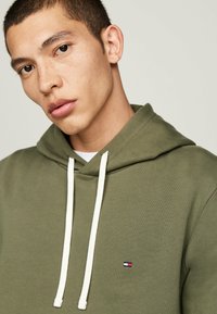 Felpa verde oliva con lacci bianchi, caratterizzata da una piccola patch con logo sul petto. Il materiale appare morbido e con una consistenza texturizzata.