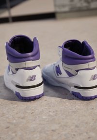 Bílé a fialové vysoké tenisky značky New Balance postavené vedle sebe na betonovém povrchu.