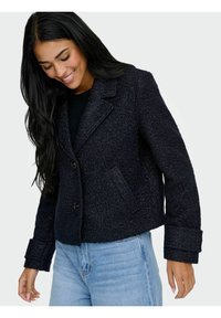 Veste courte bleu marine en tissu texturé, avec un col à revers, deux boutons et poches latérales. Associée à un jean bleu clair.