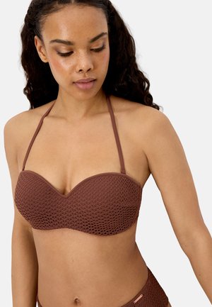 Mujer con un bikini marrón texturizado sin tirantes y con finas correas halter, mirando hacia abajo contra un fondo claro y liso.