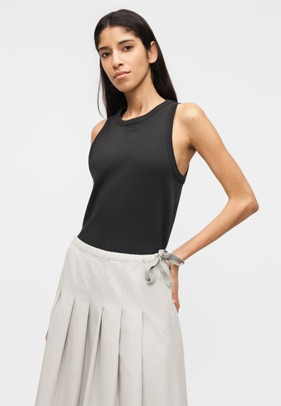 Γυναίκα που φοράει αμάνικο μαύρο τοπ και ψηλόμεση πλισέ φούστα σε ανοιχτό γκρι χρώμα δεμένη στη μέση, στέκεται μπροστά σε απλό φόντο.