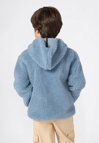 Bambino che indossa una giacca con cappuccio soffice di colore azzurro chiaro e pantaloni cargo beige, in piedi con le mani nelle tasche, rivolto di spalle su sfondo bianco.