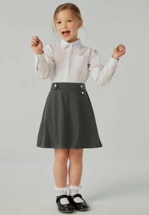 Chemise blanche à manches longues avec une poche sur la poitrine, associée à une jupe patineuse grise ornée de deux boutons blancs. Chaussures noires et chaussettes volantées.