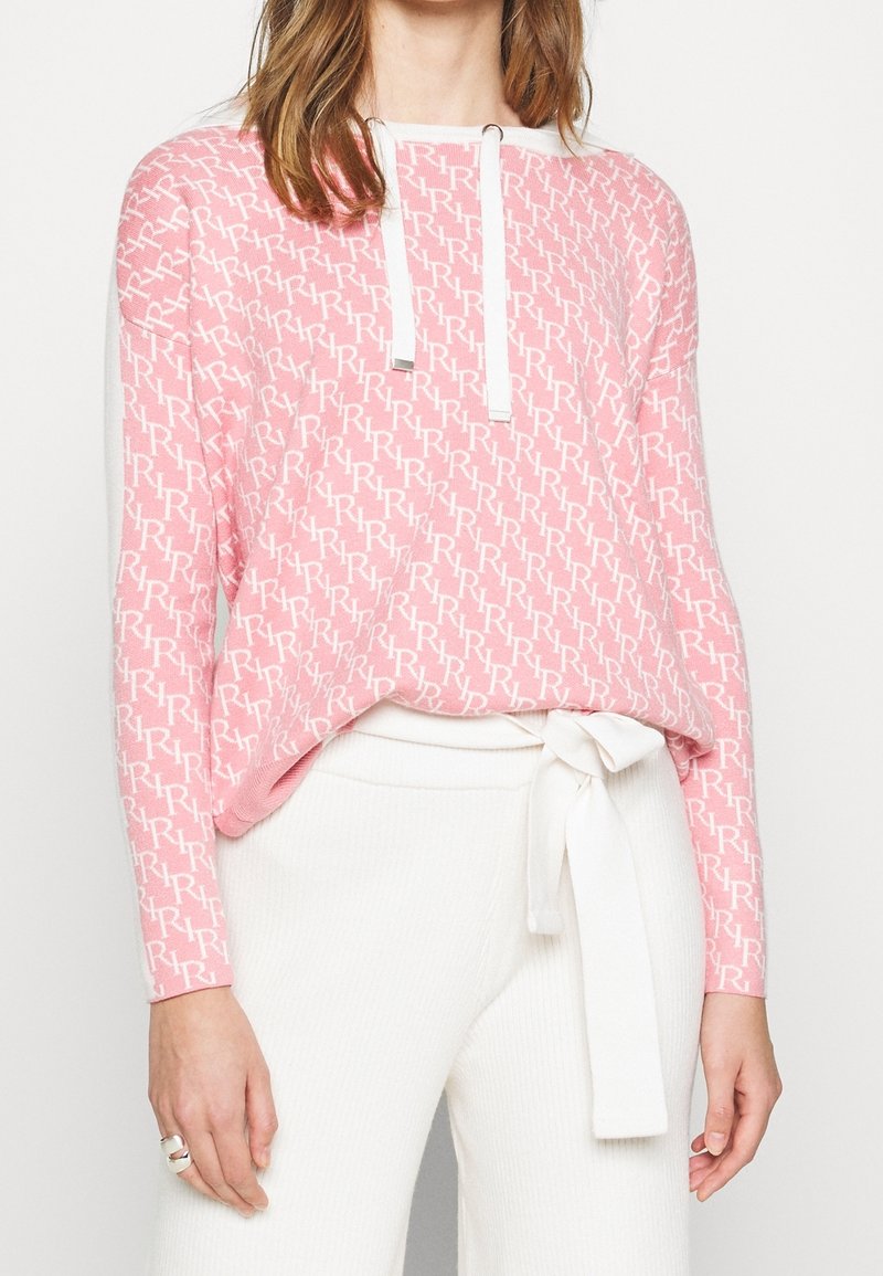 Femme portant un pull rose à motifs avec les lettres blanches « RR » et un pantalon blanc noué par un nœud à la taille, debout devant un fond uni.