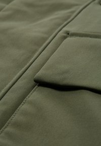 Olivová zelená tkanina s hladkou textúrou, ktorá obsahuje detail prehnaného vrecka a viditeľné švy, vytvárajúce štruktúrovaný dizajn.