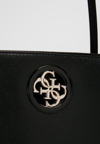 Czarna torebka o fakturze z teksturą z okrągłym logo w metalicznej złotej barwie. Projekt zawiera błyszczący czarny akcent wokół logo.