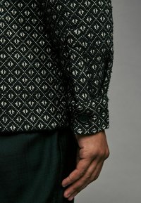 Woven black and white shirt with a diamond pattern, featuring a buttoned cuff and textured fabric, shown alongside a dark green garment.