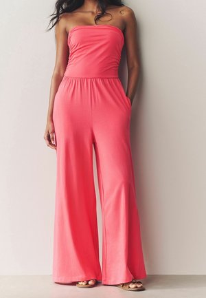 Nainen, jolla on olkaimeton korallinsävyinen haalari leveillä lahkeilla, seisoo yksivärisen seinän edessä käsivarret taskuissa ja jalassa avoimet varvassandaalit.