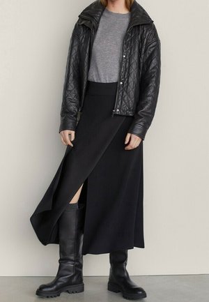 Personne vêtue d'une veste en cuir noire matelassée, d'un pull gris, d'une jupe portefeuille noire et de bottes noires montant jusqu'aux genoux, debout contre un mur uni.