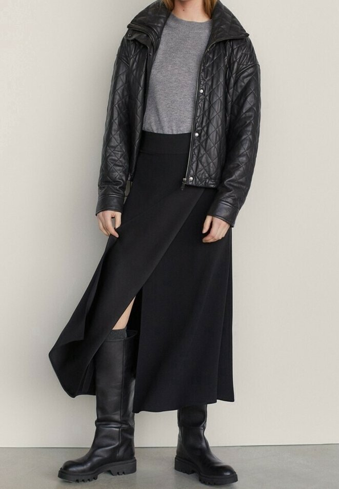 Personne vêtue d'une veste en cuir noire matelassée, d'un pull gris, d'une jupe portefeuille noire et de bottes noires montant jusqu'aux genoux, debout contre un mur uni.