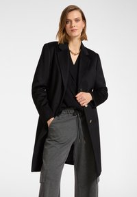 Cappotto di lana nera con design sartoriale, dotato di revers a soffietto e due bottoni frontali. Indossato sopra un top in maglia nera e pantaloni grigi.