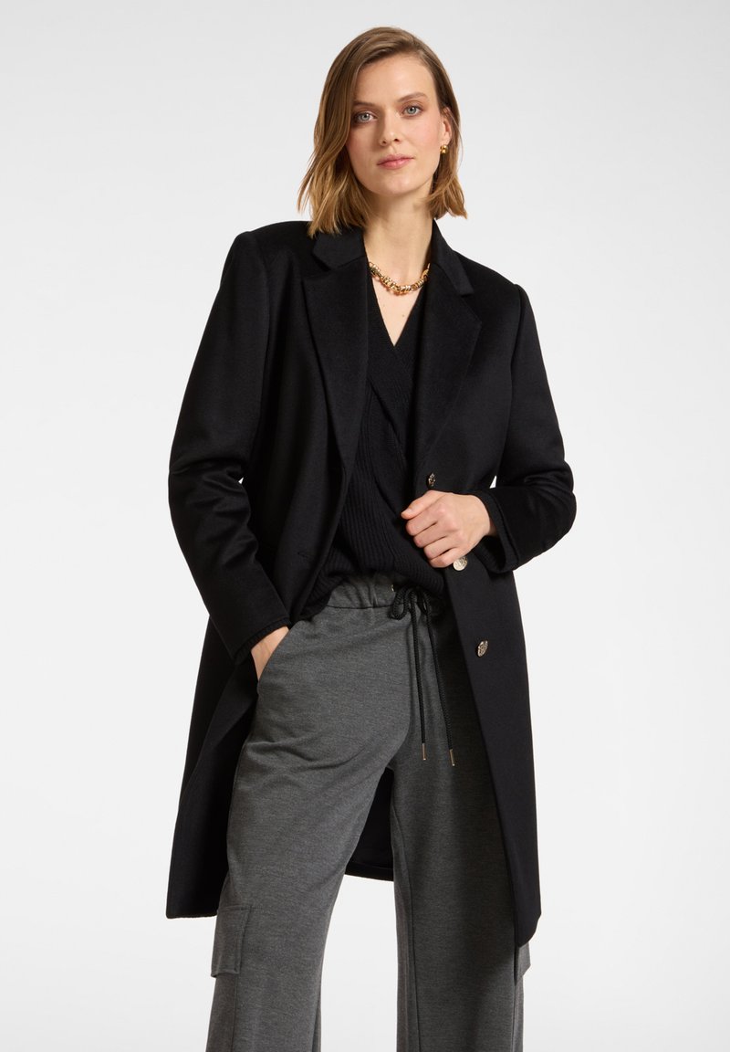 Cappotto di lana nera con design sartoriale, dotato di revers a soffietto e due bottoni frontali. Indossato sopra un top in maglia nera e pantaloni grigi.