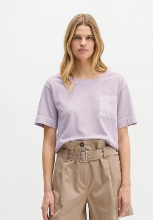 Blonde Frau trägt ein helllila T-Shirt mit kurzen Ärmeln und Tasche sowie hoch taillierte beige Shorts mit Gürtel vor einem einfarbigen Hintergrund.