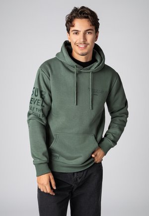 Groene hoodie met een voorzak, capuchon met koord en bedrukte tekst op de mouwen. Gecombineerd met donkere spijkerbroek, tegen een grijze achtergrond.