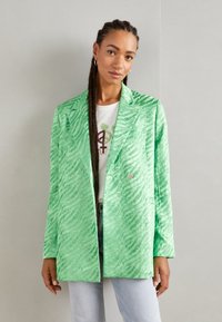 Sacou verde texturat cu un model de zebră, închidere cu un singur nasture, rever înclinat și mâneci lungi, purtat peste un tricou alb cu grafic.