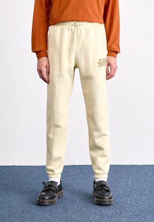 Osoba ubrana w kremowe, prążkowane spodnie dresowe z napisem „Just Do It”, w połączeniu z czarnymi mokasynami z frędzlami oraz długim rękawem w kolorze spalonym pomarańczowym.