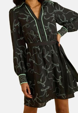 Schwarzes Kleid mit langen Ärmeln, hellgrüner Paspelierung, gepunktetem abstraktem Muster, vorne mit Knöpfen, taillierter Gürtel und gemusterte Manschetten an einer stehenden Frau.