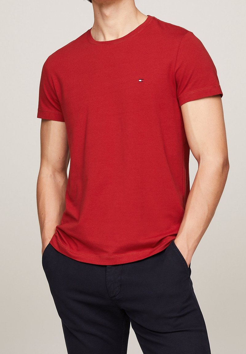 Rød bomulds T-shirt med korte ærmer, rund halsudskæring og et lille logo på brystet. Båret med mørke bukser. Glat tekstur.