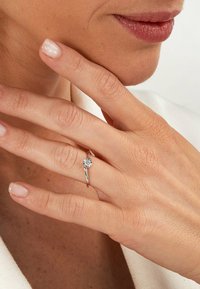 Silberner Ring mit einem quadratischen klaren Stein und einem dünnen Band. Die Hand zeigt eine natürliche Nageloberfläche, während sanftes Licht die Texturen hervorhebt.