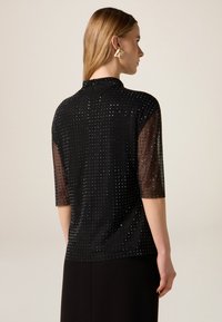 Czarna bluzka z krótkim rękawem, z jednolitym tyłem i półprzezroczystym materiałem w kropki zdobionym srebrnymi ćwiekami. Wysoki dekolt i dopasowany krój.