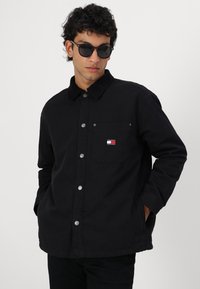 Jachetă neagră cu nasturi, cu guler din velur, buzunar frontal, capse argintii și un mic logo Tommy Jeans în roșu, alb și albastru pe piept.