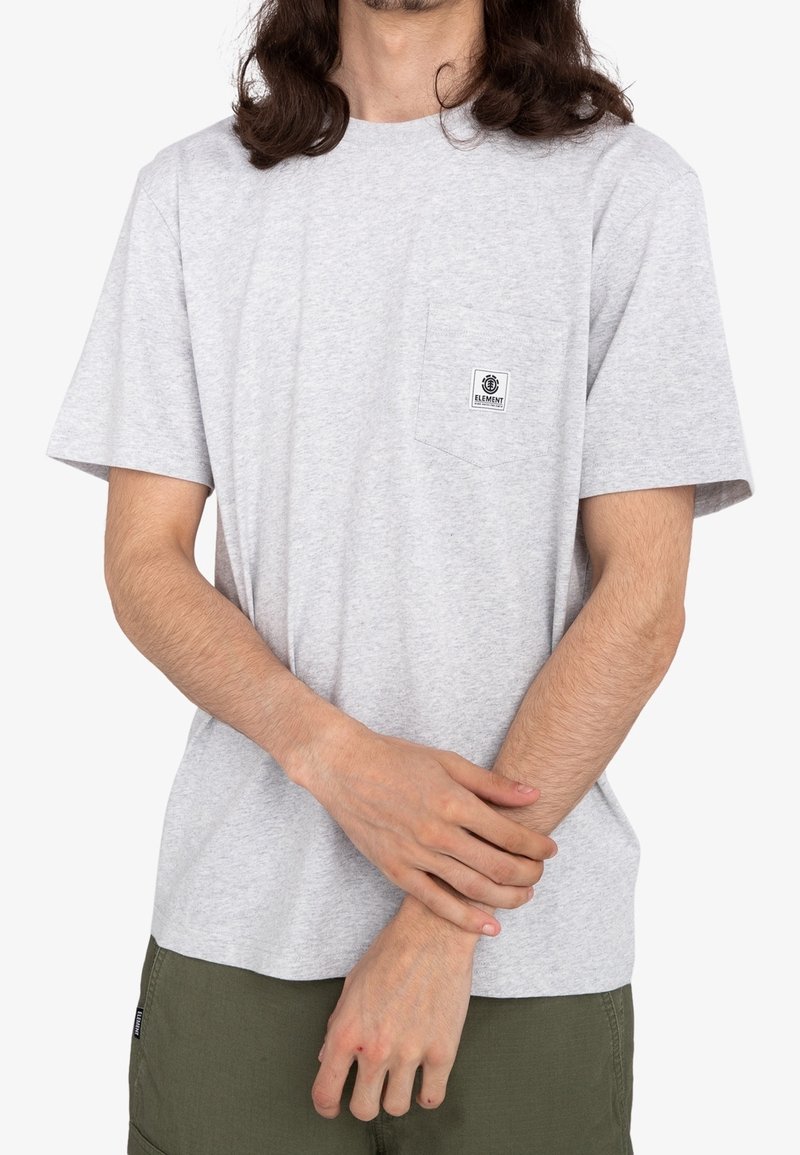 Personne aux cheveux longs portant un t-shirt gris clair avec une petite poche poitrine et un pantalon vert, mains jointes sur le poignet.