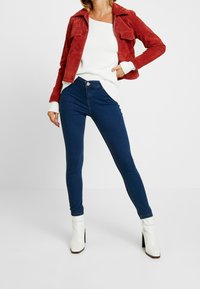 Röd mockajacka med kort modell, vit ribbad tröja, mörkblå skinny jeans och vita klackstövlar till vristerna. Fokusera på modern, åtsittande design.