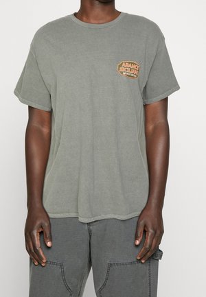 Muž má na sobě opraný šedý tričko s krátkým rukávem a oranžovým nápisem "Aranci Siciliane", které má zkombinované s tmavě šedými kalhotami s velkými kapsami.