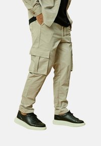 Pantalons cargo beiges avec de multiples poches et une coupe décontractée, associés à des baskets noires avec une semelle blanche et des lacets texturés.