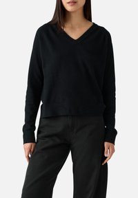 Maglione nero a V con maniche lunghe, vestibilità comoda e polsini a costine. Realizzato in tessuto morbido e liscio. Abbinato a pantaloni neri.