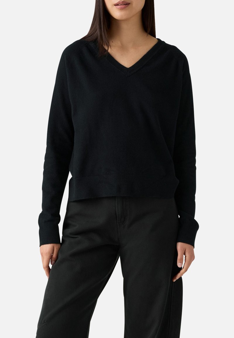 Maglione nero a V con maniche lunghe, vestibilità comoda e polsini a costine. Realizzato in tessuto morbido e liscio. Abbinato a pantaloni neri.