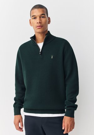 Man wearing dark green quarter-zip sweater over white shirt, standing against plain light background, facing forward with relaxed expression.