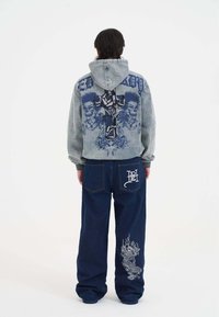 Sudadera con capucha gris que presenta un gráfico en la parte posterior de una cruz y diseños florales. Pantalones vaqueros oscuros de corte holgado con intrincados bordados en blanco de dragones.