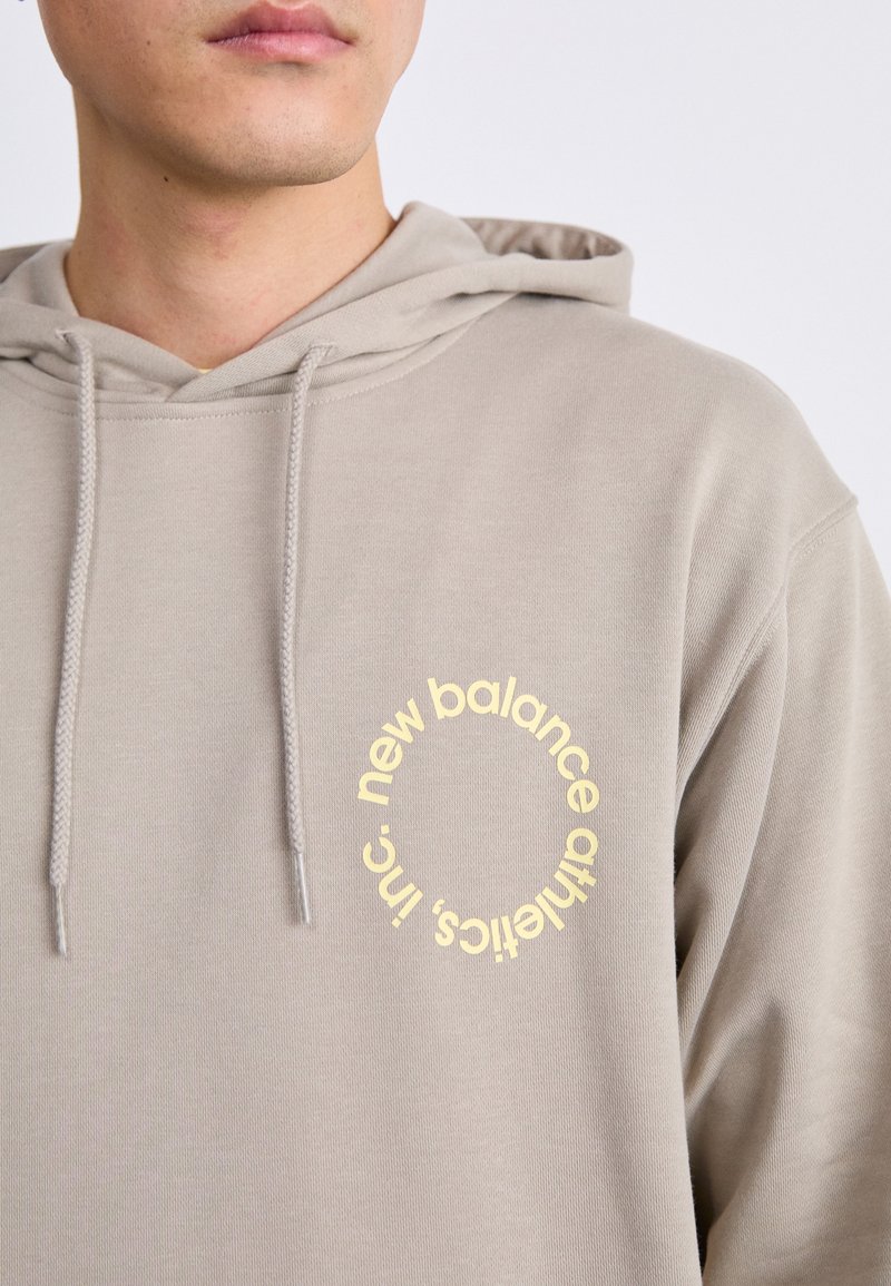 Sweat à capuche en tissu gris clair, arborant un logo circulaire jaune avec du texte sur le côté gauche de la poitrine. Texture douce avec cordons de serrage.