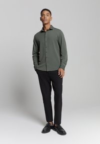 Groene longsleeve shirt met knoopsluiting,slim fit, gecombineerd met zwarte broek en zwarte loafers, tegen een neutrale achtergrond.
