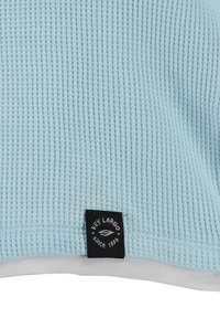 Tessuto azzurro chiaro con texture e motivo a coste, orlato di bianco; presenta un'etichetta nera con "KEY LARGO dal 1994" in lettere bianche.