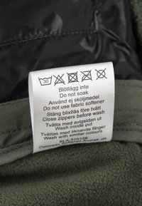 Vaskeanvisninger på plejeetiketten: vask ved 30°C, må ikke blødlægges, ingen skyllemiddel, luk lynlåse, vask vrangside ud med lignende farver.