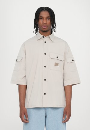 Light beige button-up shirt with short sleeves, two chest pockets, and a square patch logo, featuring black buttons and a relaxed fit.