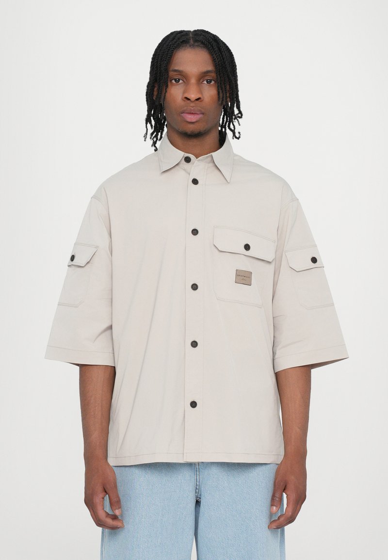 Chemise beige clair à manches courtes avec boutons, deux poches poitrine, et un logo en patch carré, dotée de boutons noirs et d'une coupe décontractée.