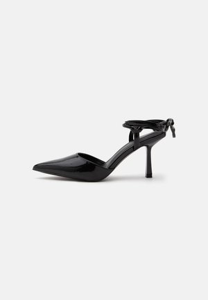 Zapato de tacón alto de cuero charol negro con punta puntiaguda y correa en el tobillo atada en un nudo en la parte trasera, mostrado sobre un fondo blanco liso.