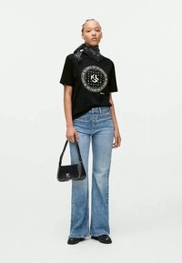 Camiseta preta com estampado gráfico, jeans azul flare, sapatos pretos planos e uma pequena bolsa preta com detalhes em prata e logotipo.
