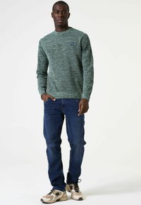 Groen gemêleerd crewneck trui, lange mouwen, gecombineerd met blauwe jeans en beige sneakers. Bevat een logo patch op de borst van de trui.